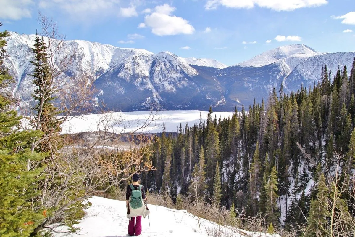 Une randonneuse descend un sentier neigeux avec vue sur des sommets enneigés et le lac Tagish gelé