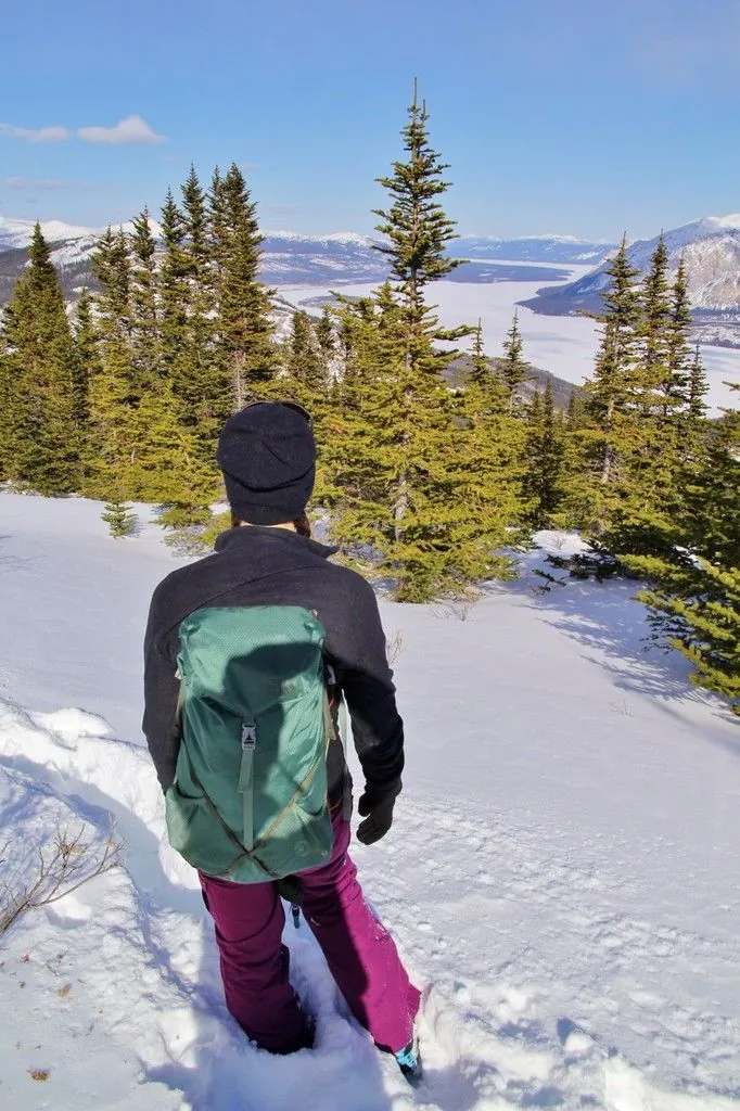 Une randonneuse au sac à dos vert de dos, enfoncée dans la neige, face au lac gelé et aux montagnes