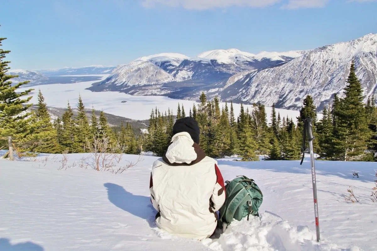 Une randonneuse assise dans la neige près de ses bâtons contemple le lac gelé et les sommets enneigés