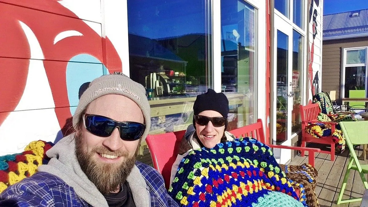 Jenni et Guigui en lunettes de soleil, couverture colorée sur les genoux, assis sur une terrasse de Carcross au soleil