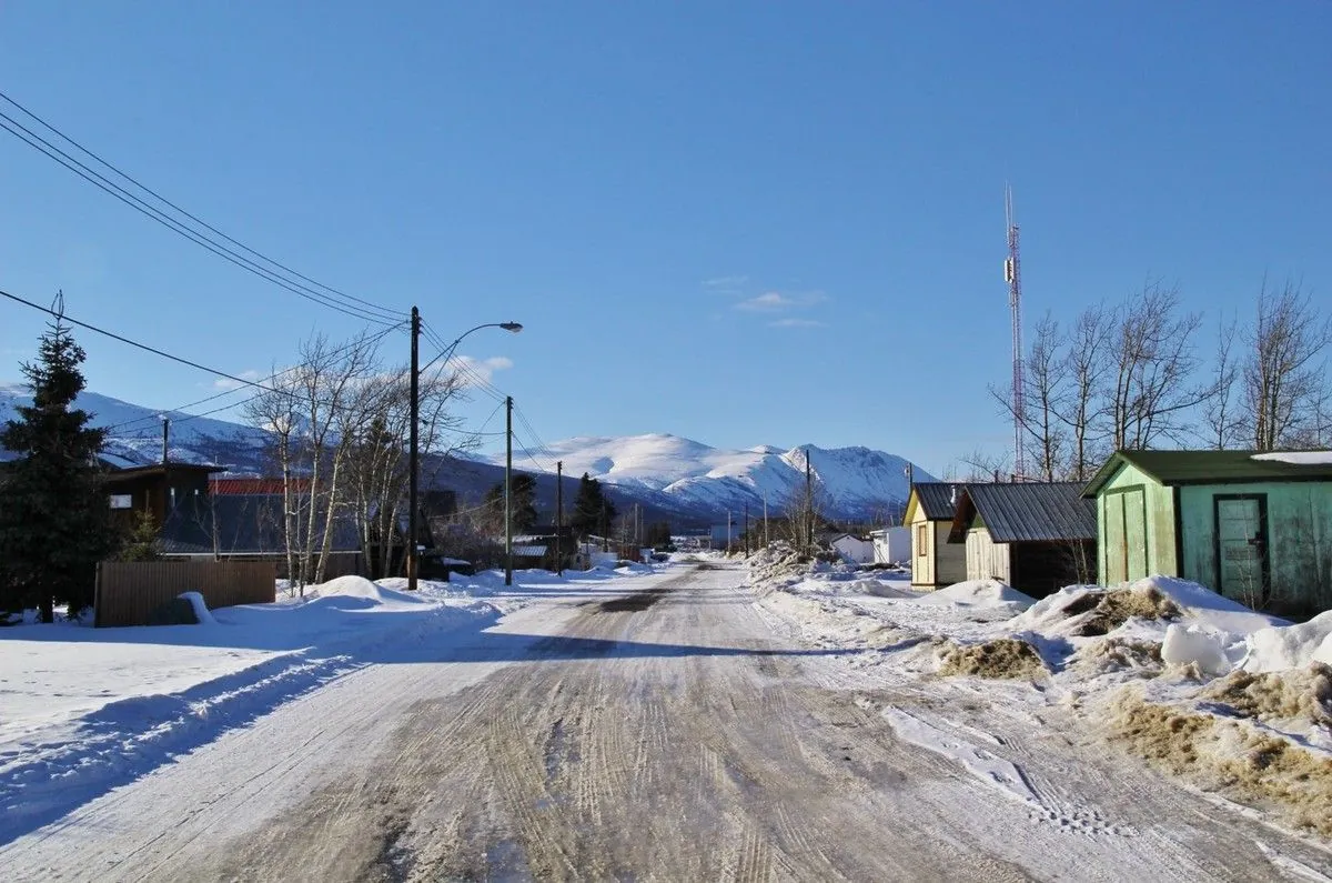 Rue déserte de Carcross, bordée de cabanes colorées, sous un ciel bleu et des montagnes enneigées