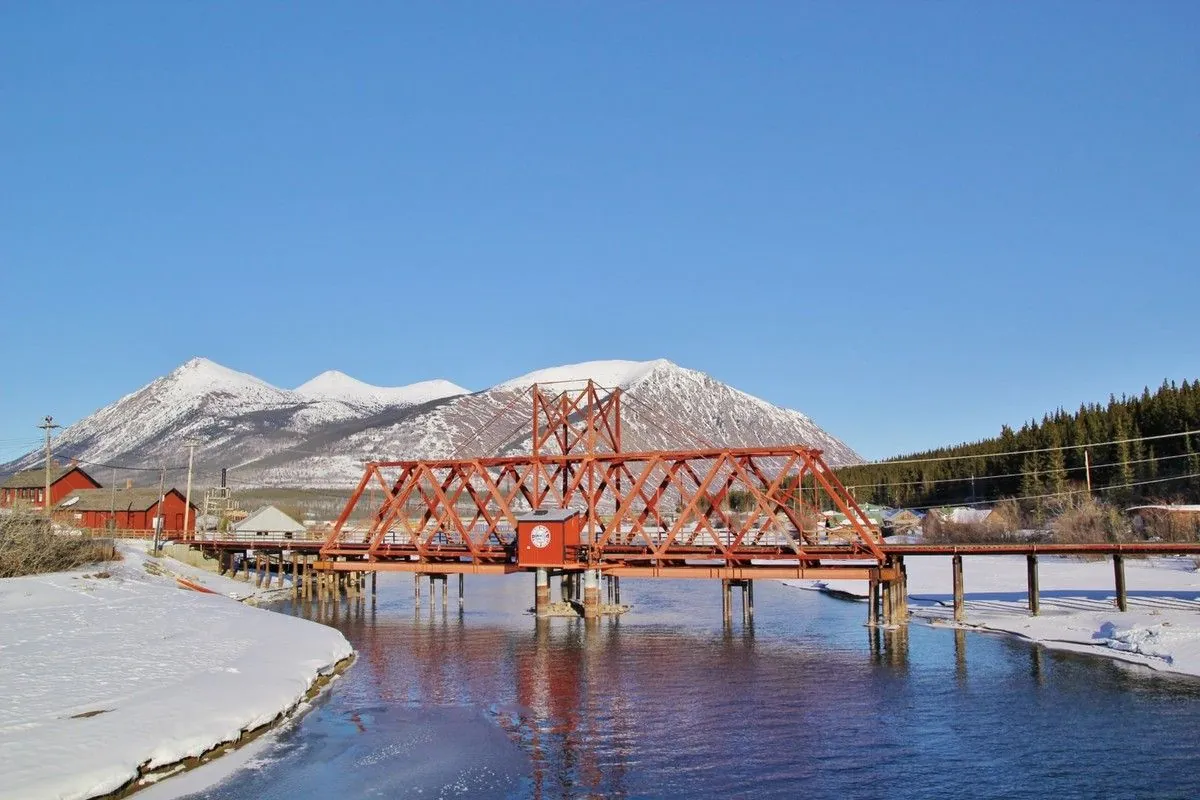 Pont ferroviaire rouge sur une eau bleue partiellement gelée, devant une montagne enneigée, à Carcross