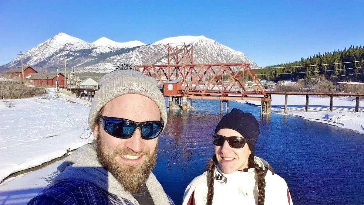 Selfie de Jenni et Guigui en lunettes de soleil devant le pont rouge de Carcross et l'eau bleue