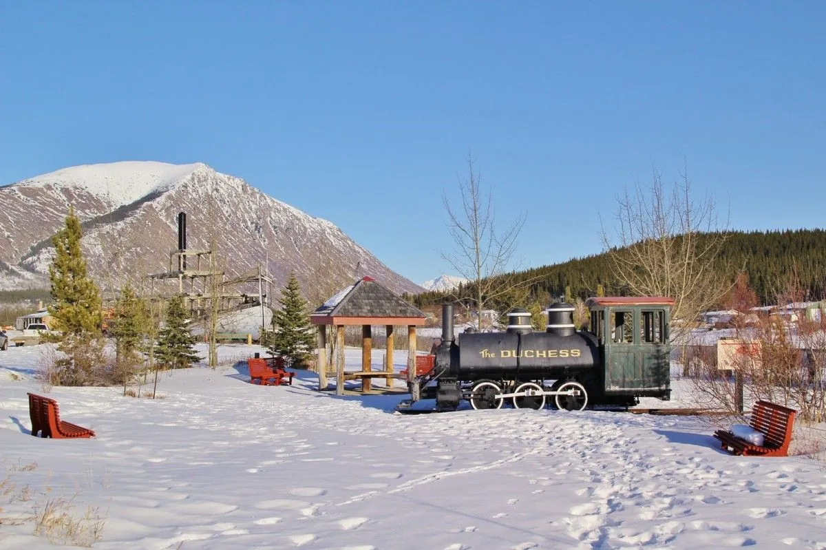 Locomotive à vapeur noire exposée dans la neige, devant des bancs rouges et une montagne, à Carcross